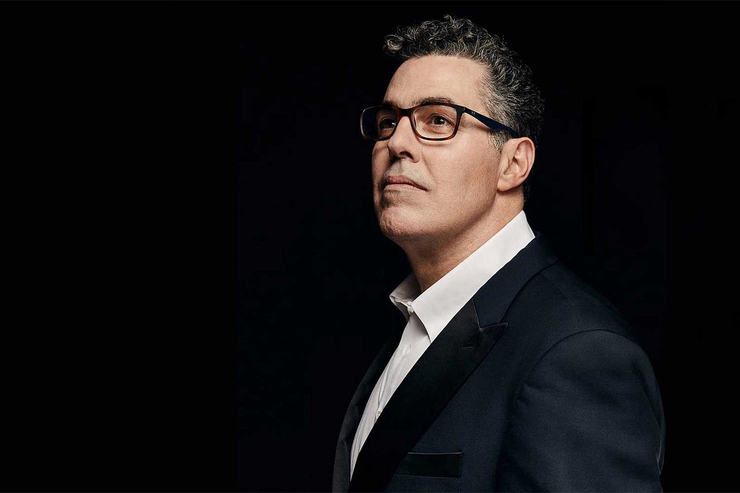 Adam Carolla in a suit looking off into the distance.