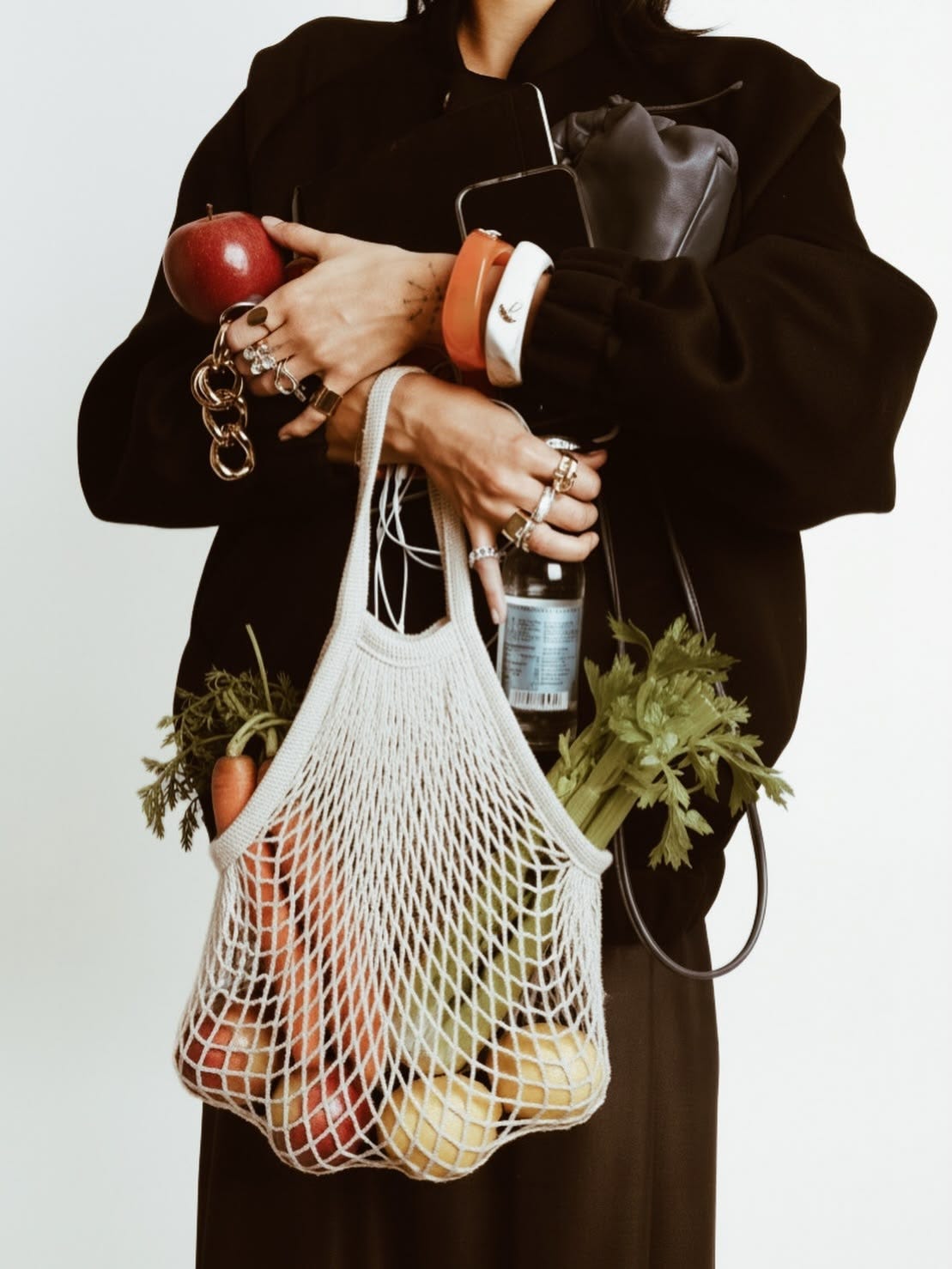 lose-up of a woman's arms carrying a net grocery bag filled with fresh produce, an apple, and a glass bottle, layered in gold jewelry lose-up of a woman's arms carrying a net grocery bag filled with fresh produce, an apple, and a glass bottle, layered in gold jewelry