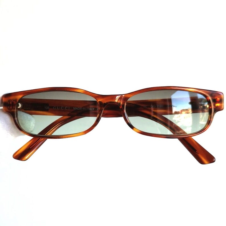 May include: Brown tortoise shell rectangular frame sunglasses with green tinted lenses. The sunglasses have the words 'GUCCI MADE IN ITALY' printed on the right lens.