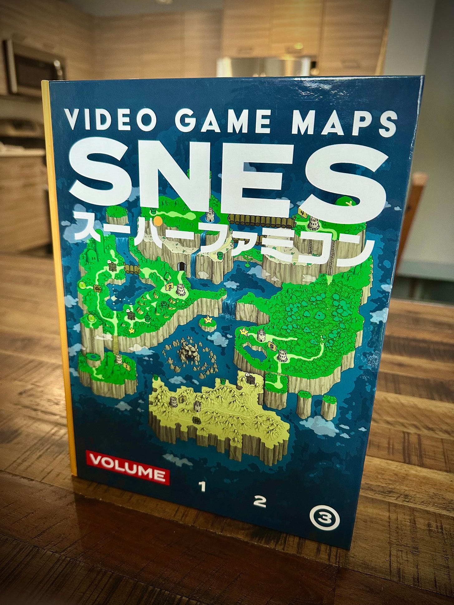 The book cover of the hardcover book video game, maps SNES volume three, which has the world map from super Mario world on the front cover a bunch of islands in the middle of an ocean that are green hills and desert, and have castles around them and stars