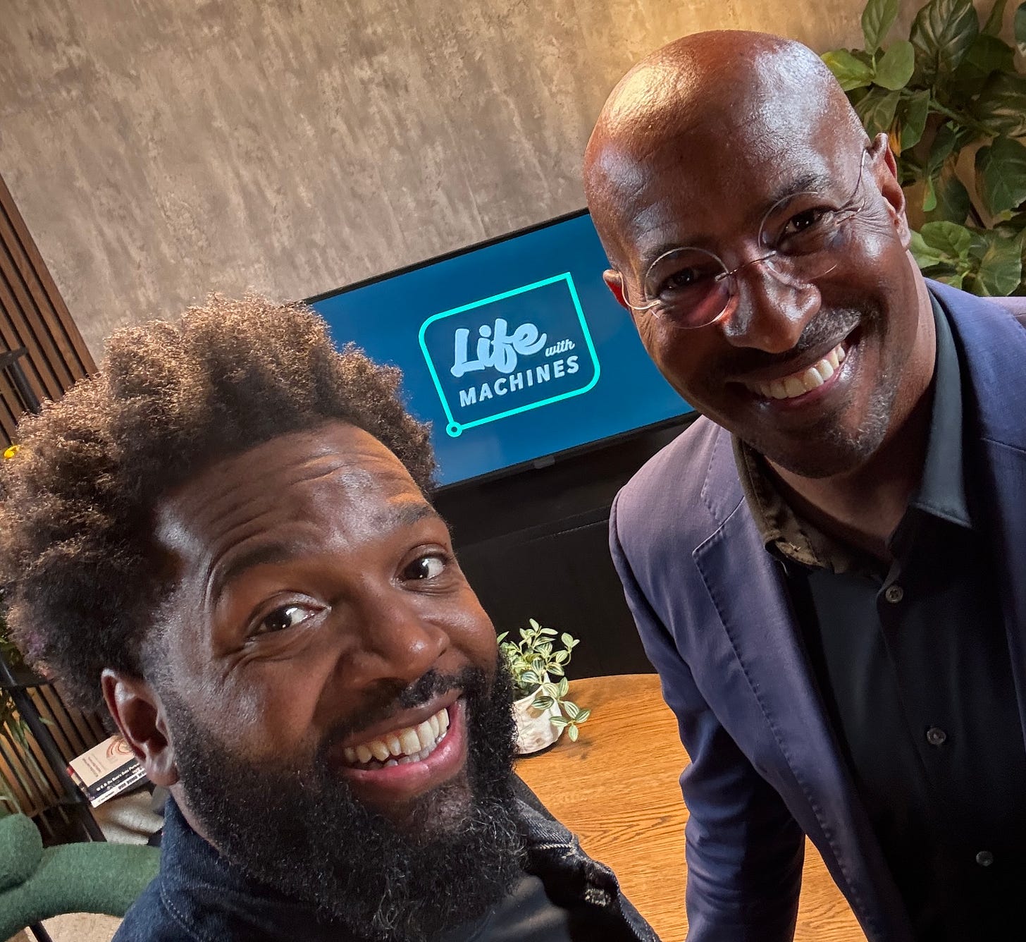 Two smiling Black men take a close-up selfie in a modern studio setting. The man on the left (Baratunde) has a full beard and natural textured hair, while the man on the right (Van Jones) is bald, wearing glasses and a navy blazer. Behind them, a screen displays the logo for ‘Life with Machines’ in a sleek, futuristic font. The setting includes a wooden table, green plants, and soft ambient lighting, creating a warm and professional atmosphere.