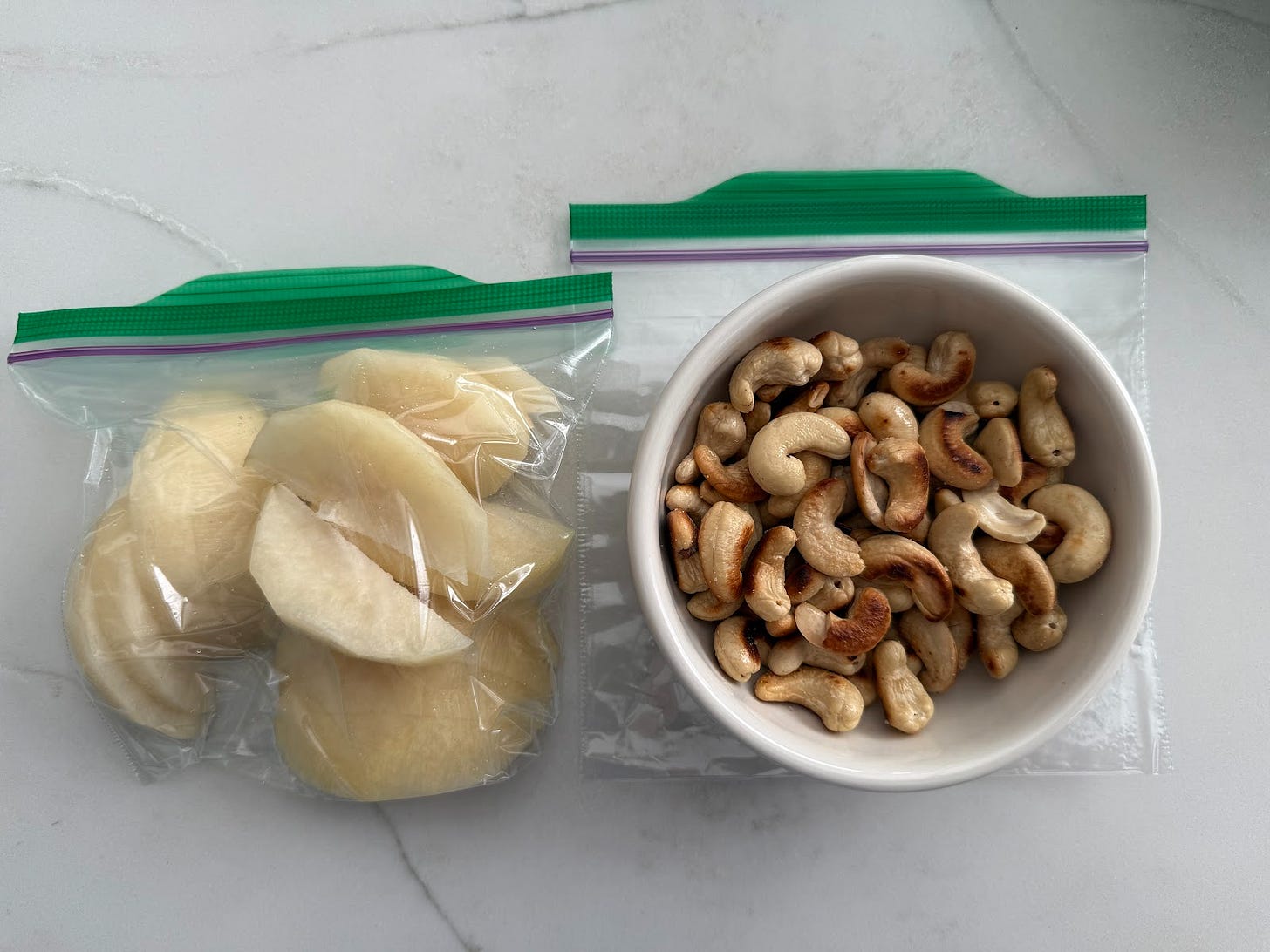 A small bowl of roasted cashews and a plastic bag of sliced Asian pear, both placed neatly on a white surface. A small bowl of roasted cashews and a plastic bag of sliced Asian pear, both placed neatly on a white surface.