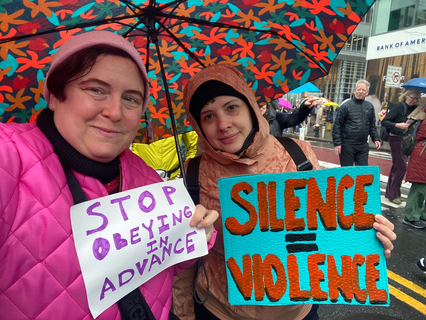Two women hold signs that read: Stop Obeying in Advance and Silence = Violence