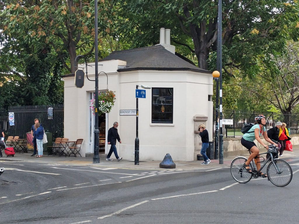 A photo of WatchHouse coffee shop, in an old watch house in Bermondsey, London. Pedestrians and a bicyclist move past. A photo of WatchHouse coffee shop, in an old watch house in Bermondsey, London. Pedestrians and a bicyclist move past.