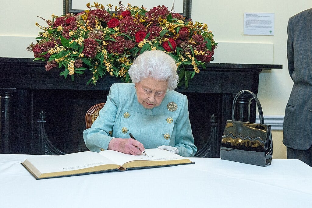 A photo of Queen Elizabeth II signing an official book with her handbag next to her on the table.