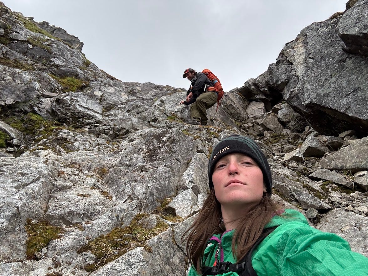 A phone selfie taken mid-descent: a woman in a black beanie and green rain jacket looks into the camera with a flat, determined expression, filling the lower right of the frame. Behind and above her, a man in a grey jacket, cap, and orange pack picks his way down a gully of wet grey rock under a flat white sky. A phone selfie taken mid-descent: a woman in a black beanie and green rain jacket looks into the camera with a flat, determined expression, filling the lower right of the frame. Behind and above her, a man in a grey jacket, cap, and orange pack picks his way down a gully of wet grey rock under a flat white sky.