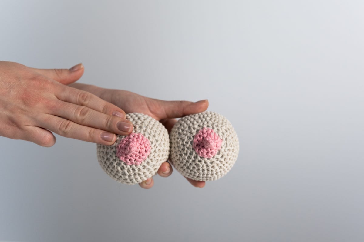Hands holding two white cotton balls with pink “nipples”-they look like breasts.