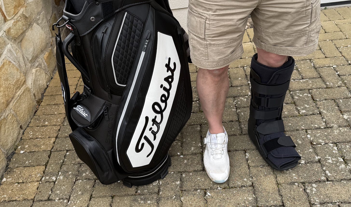 A golfer standing beside a golf bag, wearing a white golf shoe on one foot and a black medical walking boot on the other.