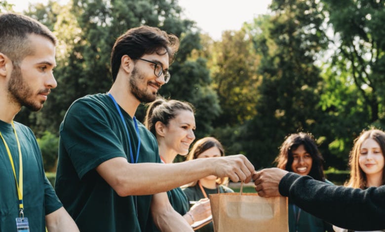 A group of volunteers in green shirts and name badges handing a bag of food to a recipient at an outdoor food distribution event, with lush greenery visible in the background.