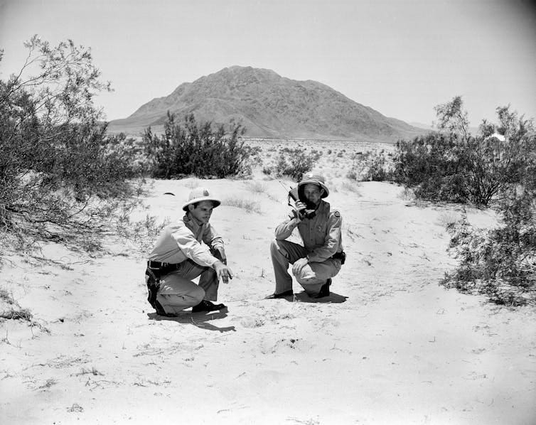 Two men wearing hats crouch on the ground in the desert, with bushes and a mountain around them. Two men wearing hats crouch on the ground in the desert, with bushes and a mountain around them.