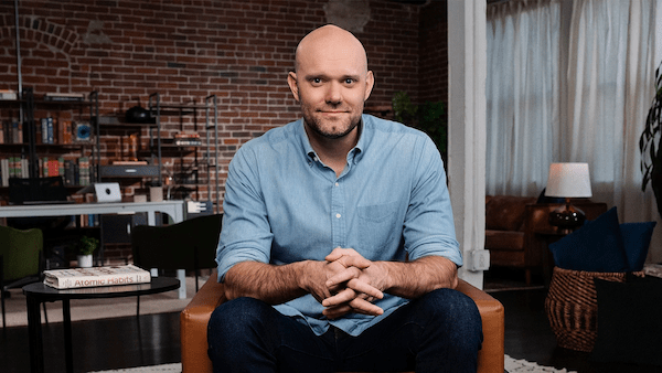 A man with a shaved head wearing a light blue button-up shirt is sitting on a brown chair, looking directly at the camera. The background features a brick wall, a bookshelf, and a softly lit room with plants and furniture. A man with a shaved head wearing a light blue button-up shirt is sitting on a brown chair, looking directly at the camera. The background features a brick wall, a bookshelf, and a softly lit room with plants and furniture.