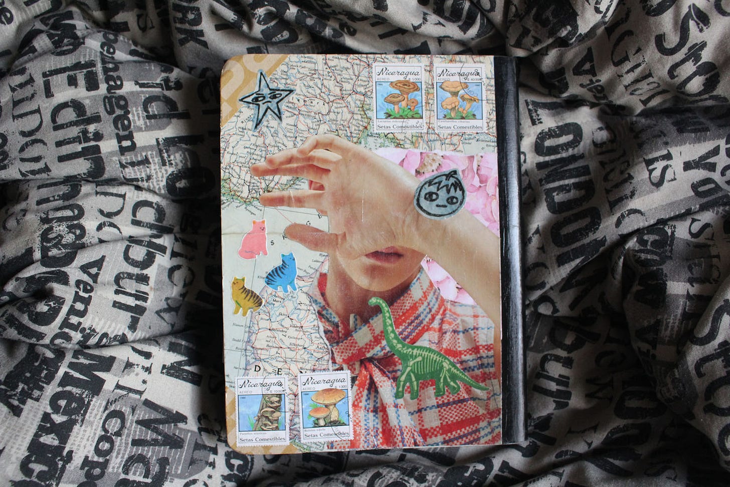 the back of a colorfully collaged composition book with a picture of a person covering their face, cat stickers, stamps, and a map