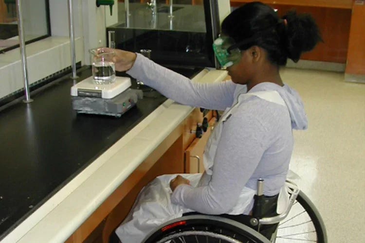 Person in a wheelchair sitting at a table in a lab, measuring the weight of a beaker Person in a wheelchair sitting at a table in a lab, measuring the weight of a beaker