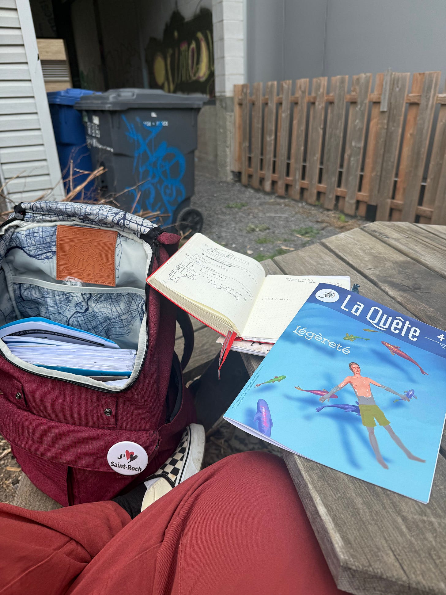 A backyard wooden table with an open notebook and a copy of La Quête magazine, cover reading "Légèreté," beside a burgundy backpack bearing a J'aime Saint-Roch button. Checkered sneakers visible. Recycling bins with graffiti in the background.