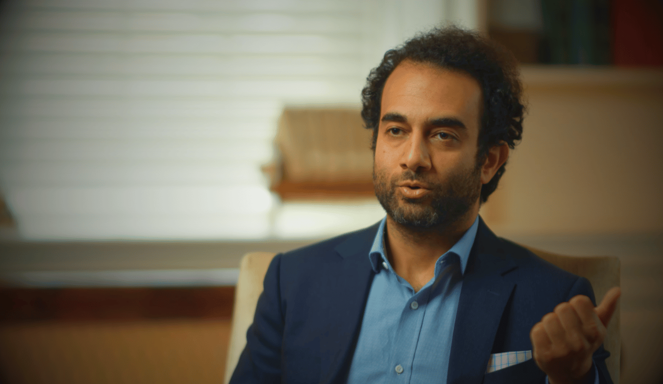 Shadi Hamid is a middle-aged man of Middle Eastern appearance in a blue suit speaking while seated indoors with blurred bookshelves in the background.