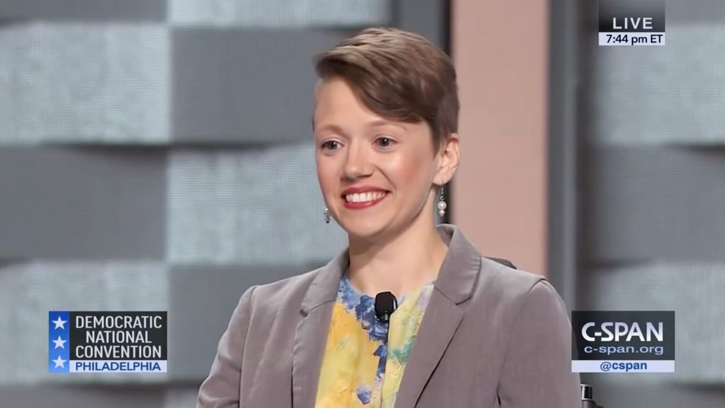 A screenshot of a white disabled person with short hair wearing a blazer and watercolor-like shirt and dangly earrings, smiling widely. Text on the image shows it is from a C-SPAN broadcast of the Democratic National Convention in Philadelphia. Live. 7:44pm ET.