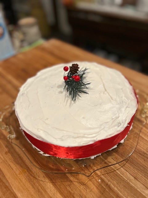 A white cake with a red ribbon ted around it and sprig of pine as the centerpiece; an empty pie shell with cookie cutters and scraps of pie crust on a wooden cutting board.