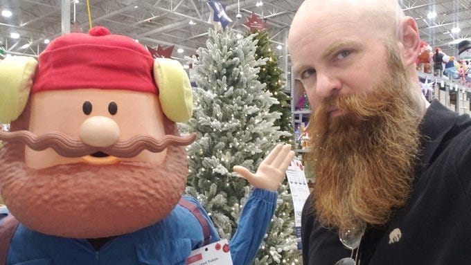 A bald man with a long red beard wearing a black hoodie and glasses stands in a store aisle next to a large plush white character with a round body black mustache and wide eyes dressed in a red knit hat with yellow earflaps and blue overalls gesturing with one hand raised the background includes a small artificial Christmas tree with white decorations hanging lights and shelves of holiday merchandise. A bald man with a long red beard wearing a black hoodie and glasses stands in a store aisle next to a large plush white character with a round body black mustache and wide eyes dressed in a red knit hat with yellow earflaps and blue overalls gesturing with one hand raised the background includes a small artificial Christmas tree with white decorations hanging lights and shelves of holiday merchandise.