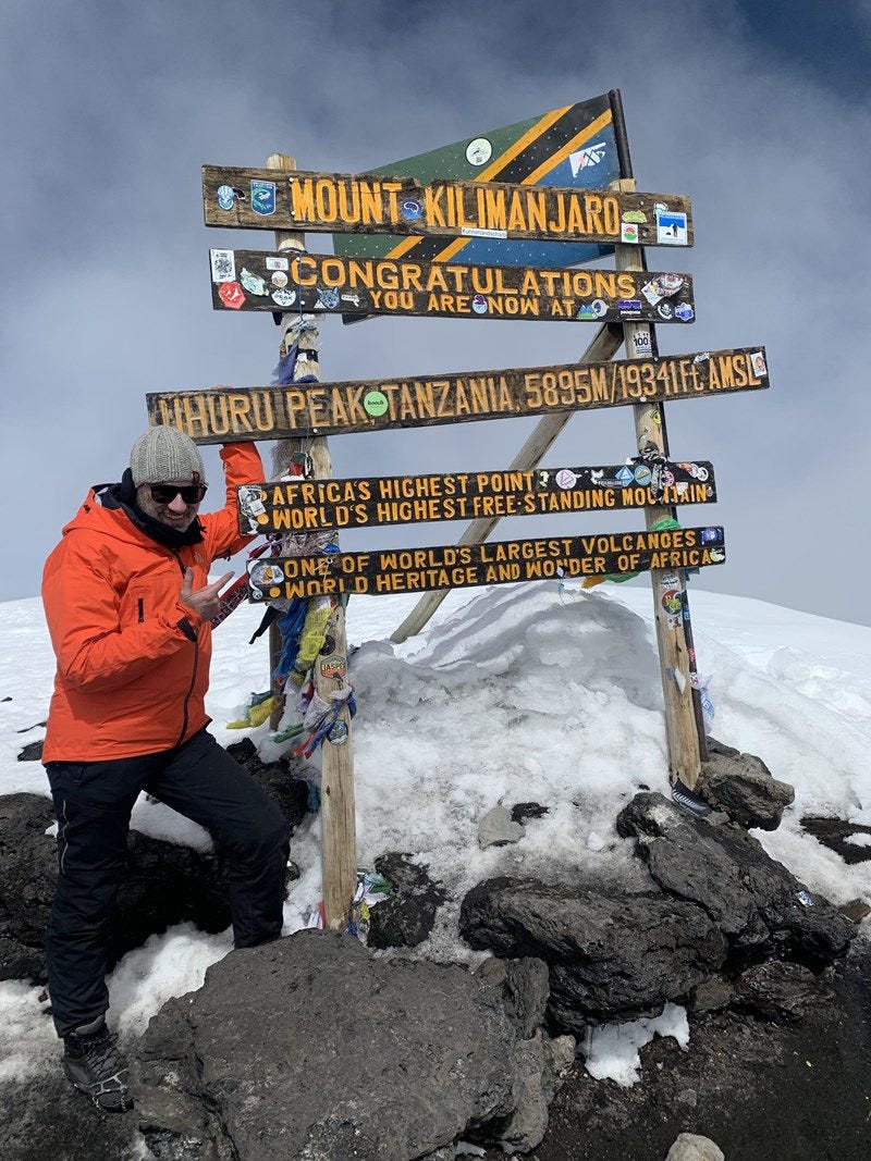 David is standing on large rocks, covered in snow, next to a sign that reads "Mount Kilimanjaro, congratulations"