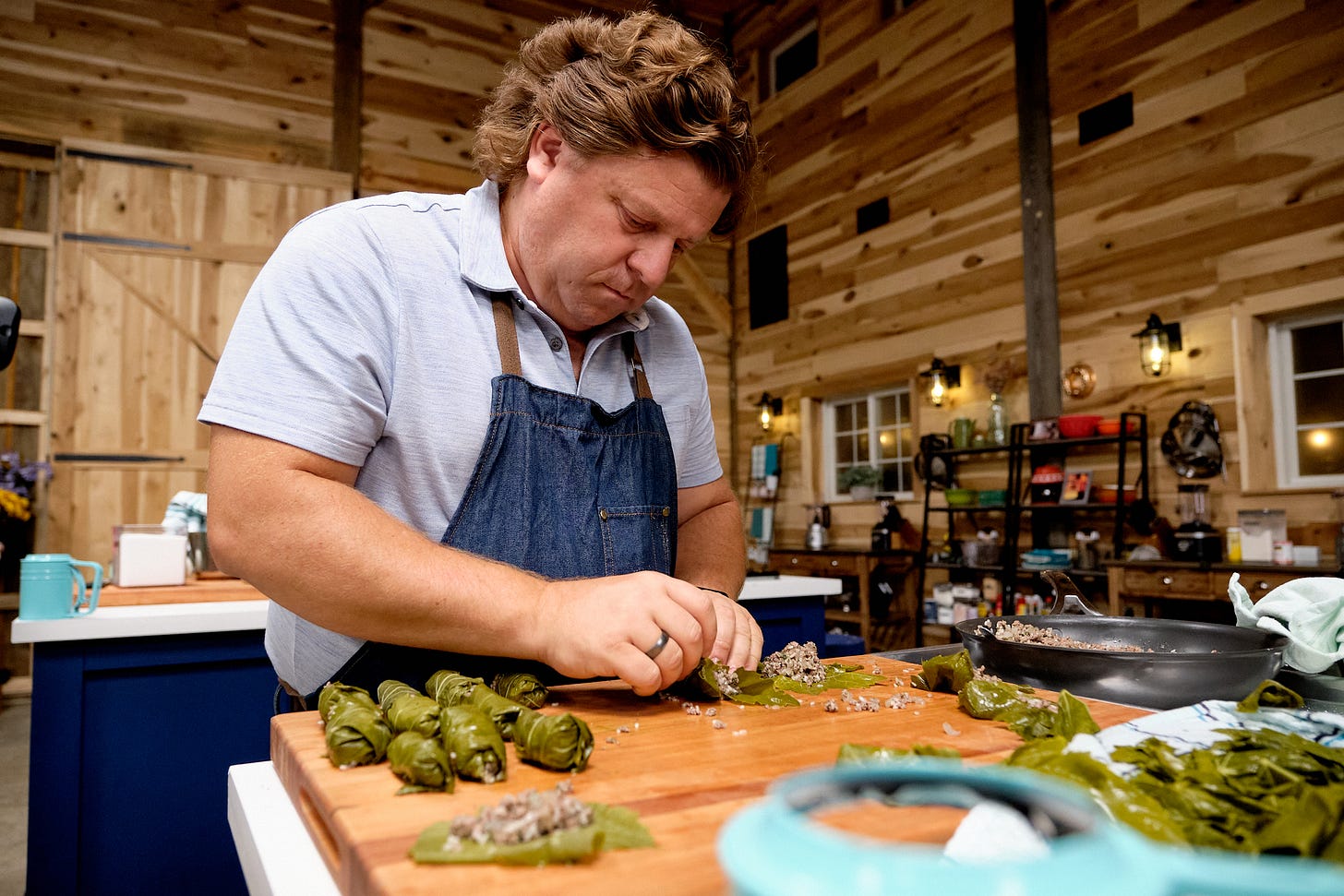 Kitchen setting, a white man stands behind a cutting board prepping stuffed grape leaves