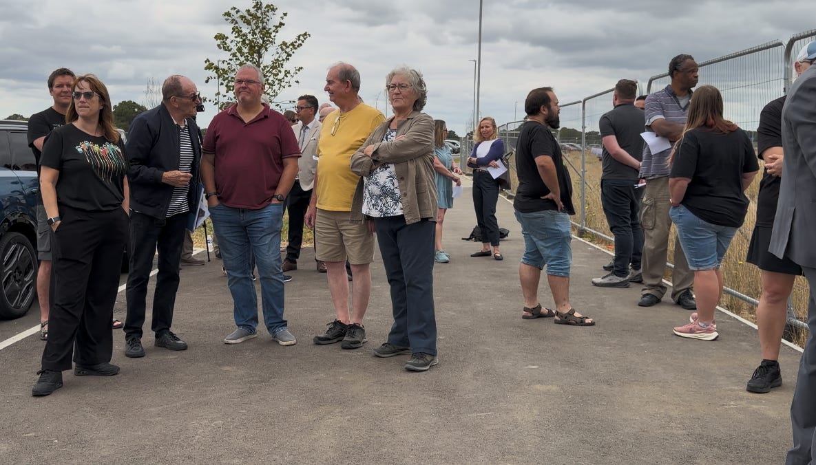 A group of adults of different ages stand on a pavement beside a harris fenced section of land under a cloudy sky. Some hold campaign papers while others look on whilst gathering for the Chilmington Secondary School Transport for Chilmington meeting begins. Parked cars line the left side, and behind the fence are grassy and dry patches with a few trees in the distance.