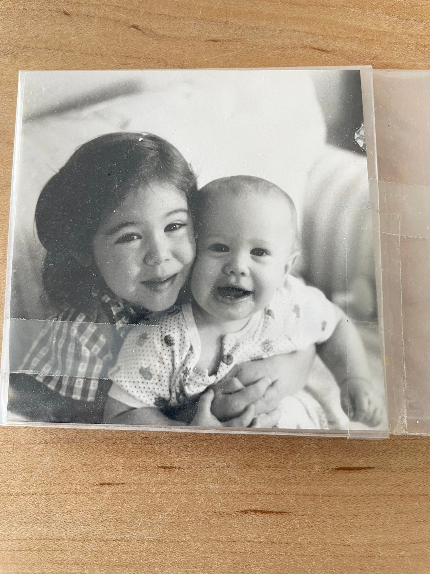a black and white photo of a four year old girl hugging her infant brother, on a maple table. a black and white photo of a four year old girl hugging her infant brother, on a maple table.
