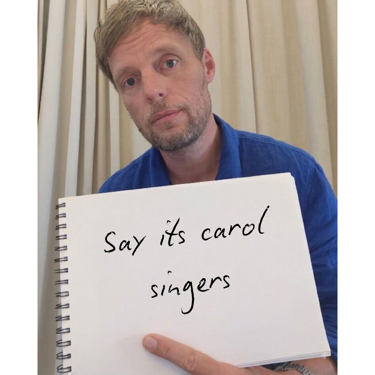 Man in a blue shirt holds up a spiral notebook to the camera, Love Actually cue card style. Handwritten: "Say its carol singers"