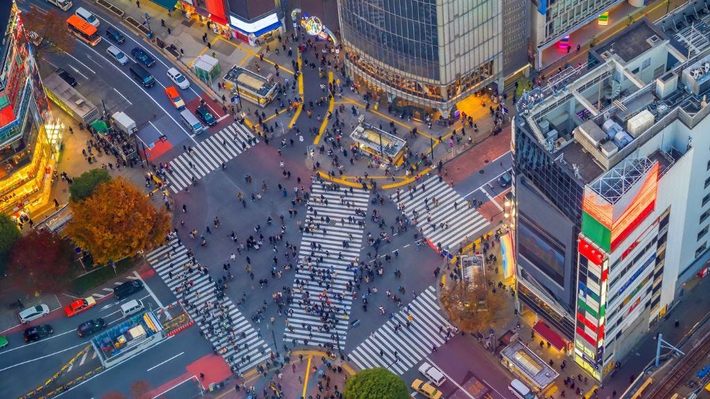 Aerial view of Tokyo’s bustling Shibuya Crossing, with crowds of people walking in all directions across the iconic diagonal crosswalks surrounded by tall buildings and traffic.