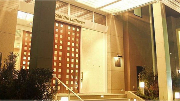 A collage of four photos of Hotel the Lutheran in Osaka. The top left shows the modern exterior entrance lit warmly at night. The top right shows a bright, minimalist guest room with a double bed, wooden floors, and simple furnishings. The bottom left highlights a wide hallway with handrails along the wall, suitable for accessibility needs. The bottom right shows a spacious accessible bathroom with grab bars, a roll-in shower with a curtain, and a toilet designed for ease of use.