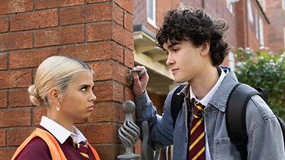 A girl and boy stand together by a wall looking tense