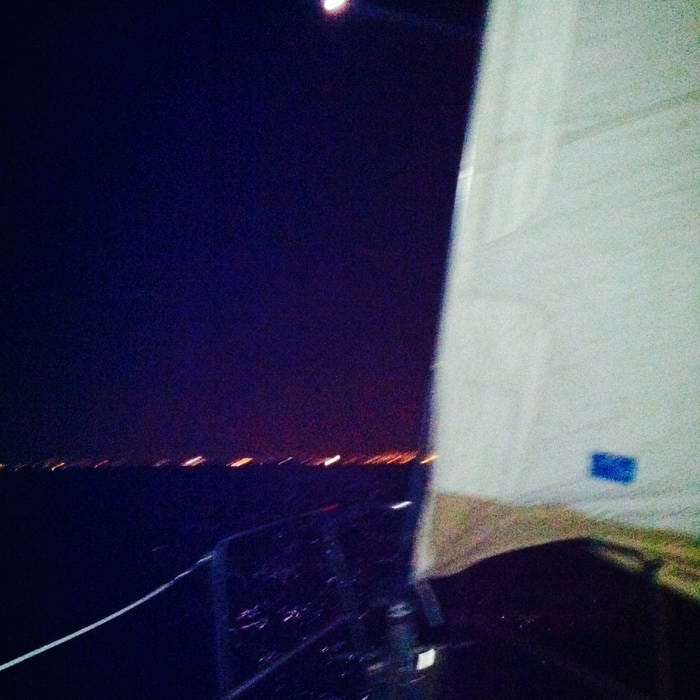 a view of Lake Michigan from a sailboat at night. The lights of Chicago are visible in the background, a sail takes up the right third of the frame. The night is indigo and watery