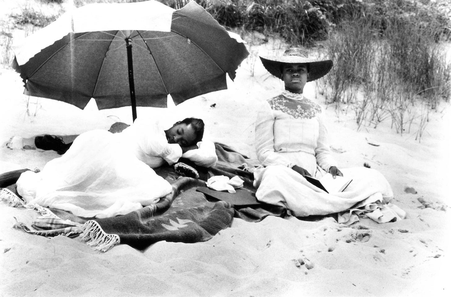 Malindy on a beach with her friend who is asleep on the blanket under an umbrella. 