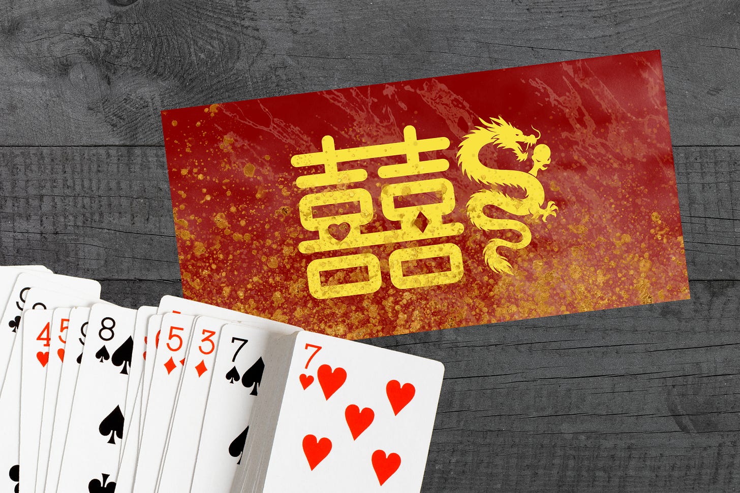 A top-down view of a red Chinese envelope (hongbao) resting on a dark grey wooden surface. The envelope features gold speckles, a dragon illustration, and the large golden "Double Happiness" character (囍). Below it lies a spread of playing cards, with the Seven of Hearts visible on the face of the packet.