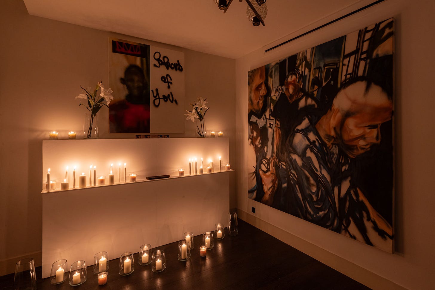 Low-lit room corner with rows of lit candles along the floor and a wall shelf, creating a warm, devotional atmosphere around the artwork.