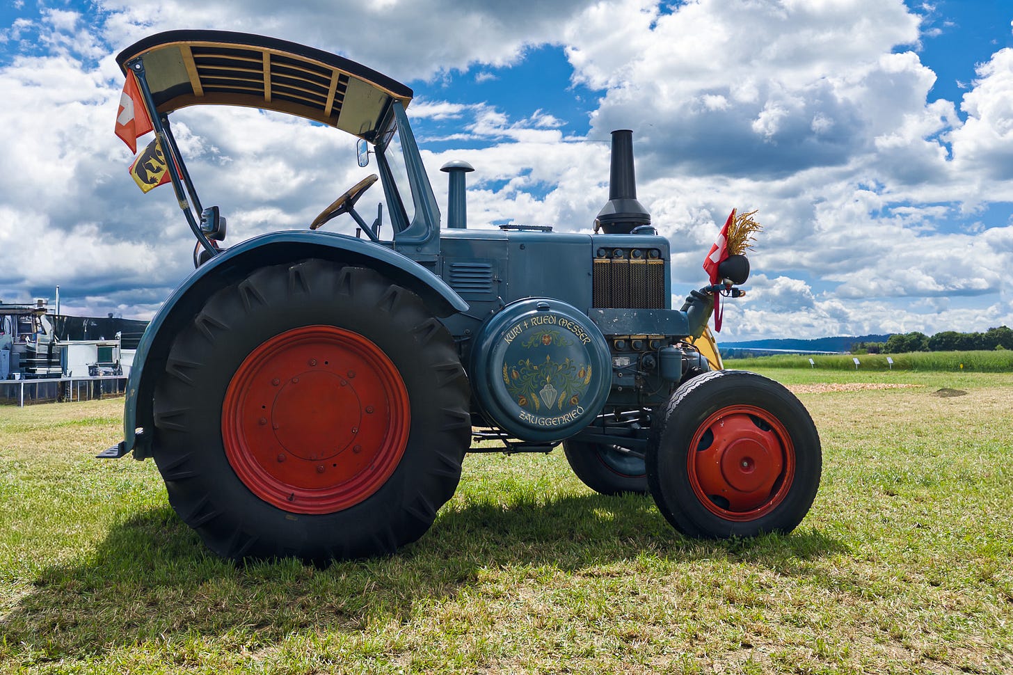 Ein Lanz Bulldog D 9506 Oldtimer steht auf einer gemähten Wiese. Der Traktor ist grau-blau lackiert, hat grosse schwarze Reifen mit roten Felgen, ein schlichtes Sonnendach und die typische markante Front historischer Lanz-Modelle. Am Heck wehen eine Schweizer und eine Berner Fahne, vorne am Kühler ist dekorativ ein kleines Bündel Getreide angebracht. Im Hintergrund öffnen sich Felder und ein weiter Himmel mit grossen Quellwolken.