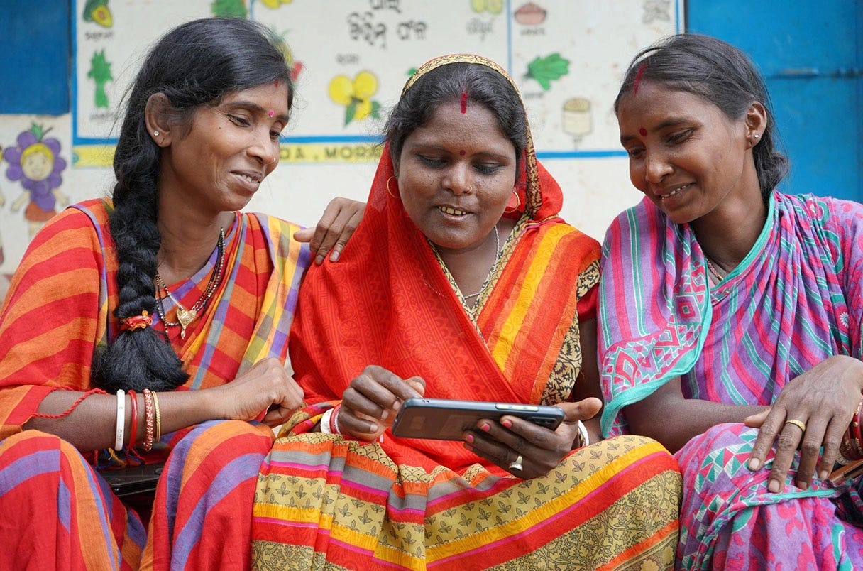 Three Indian women sitting together with their attention focused on something on a phone screen in front of the middle woman. Three Indian women sitting together with their attention focused on something on a phone screen in front of the middle woman.