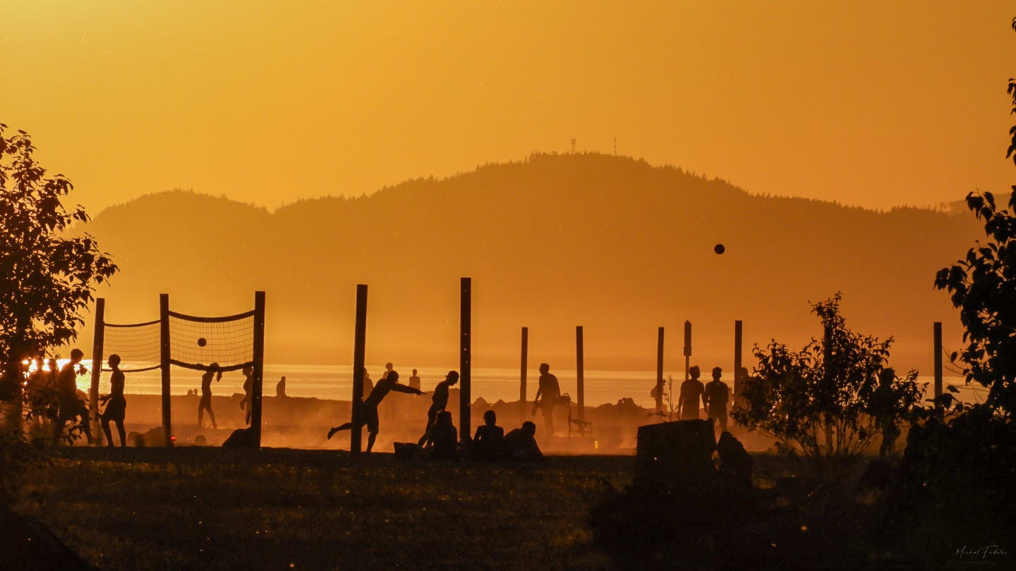 Young people playing volleyball on a beach against the setting sun, with mountains in the background.