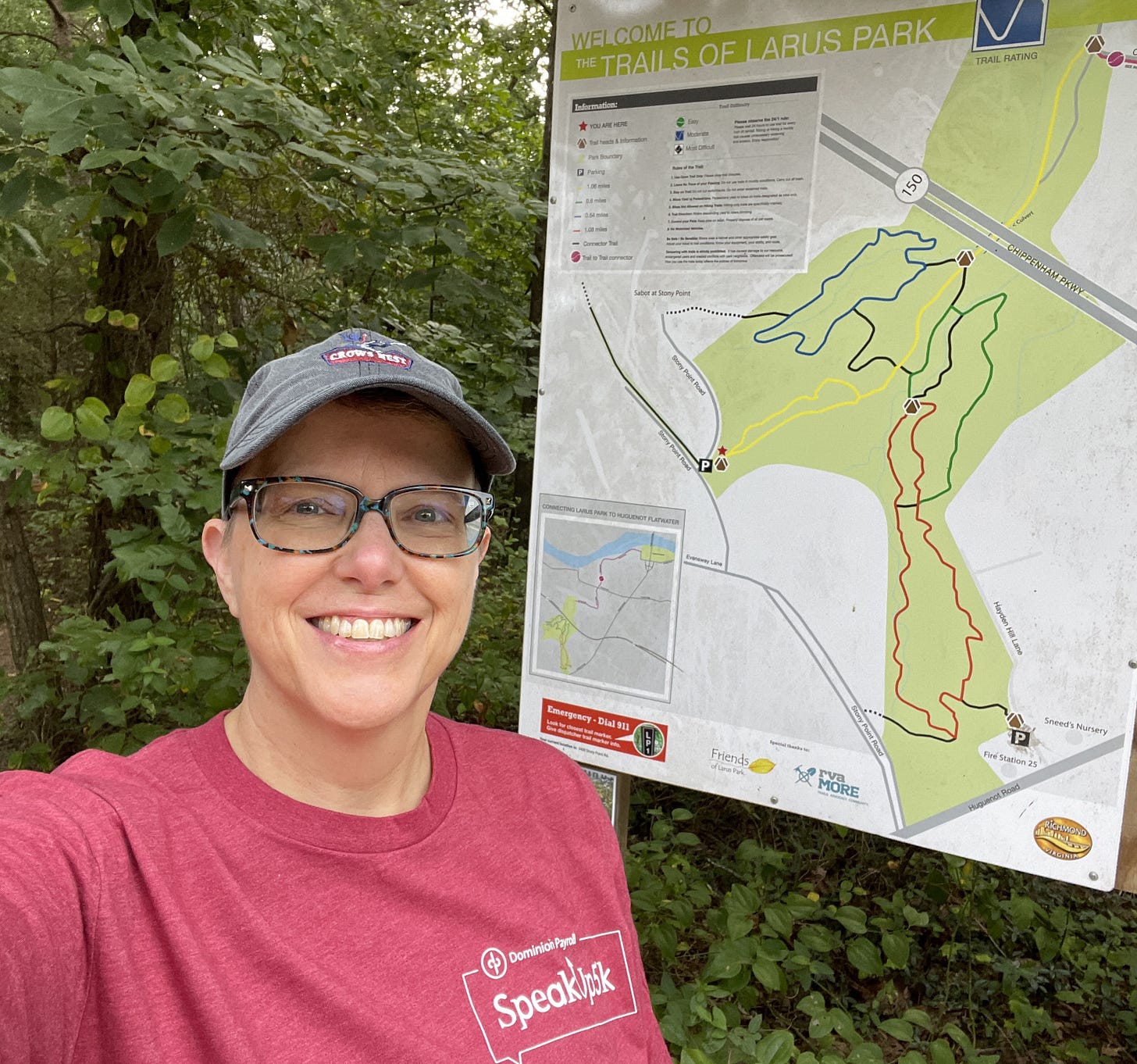 white woman wearing glasses, ballcap, and red-tshirt smiling at camera in front of map of trails.
