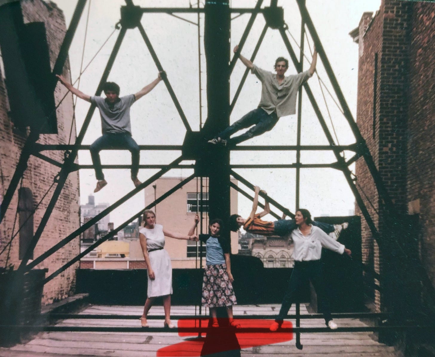 People hanging off the metal braces of a rooftop water tower between two brick walls; a girl is centered with red marking highlighting the tar roof beneath her.