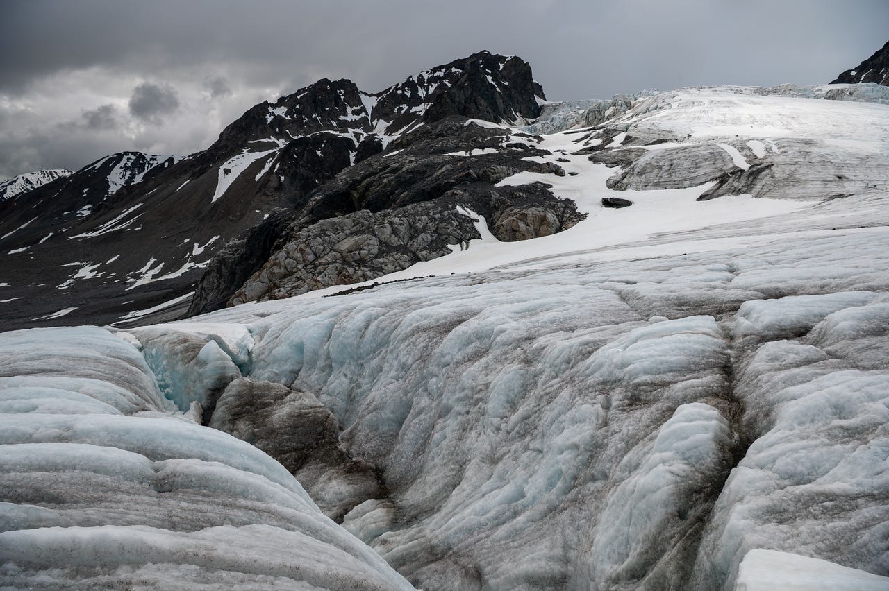 Standing low on the buckled ice of an icefall, looking across ridges of dirty grey-blue glacier ice split by crevasses, one dark slot opening just right of center. The ice rises in pressure ridges toward pale blue seracs above, and beyond them a black rock peak with snow-filled gullies stands against a flat grey sky. Bands of rock dust streak and darken the ice everywhere except a few clean white patches of snow.