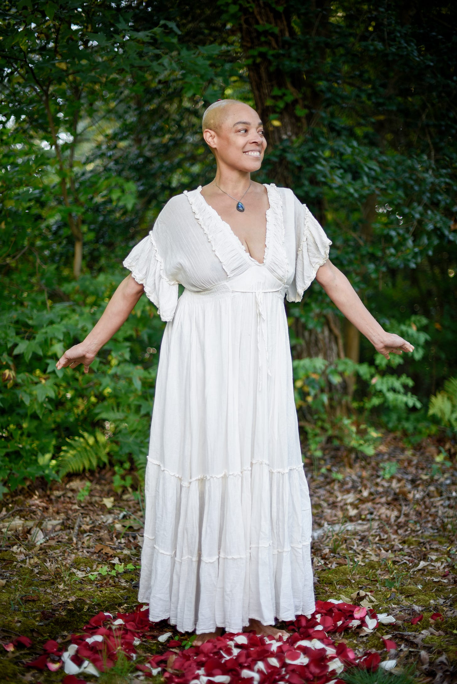 Doc Jana aka Radiance standing happily in the woods wearing a flowy white dress with red and white rose petals encircling their feet