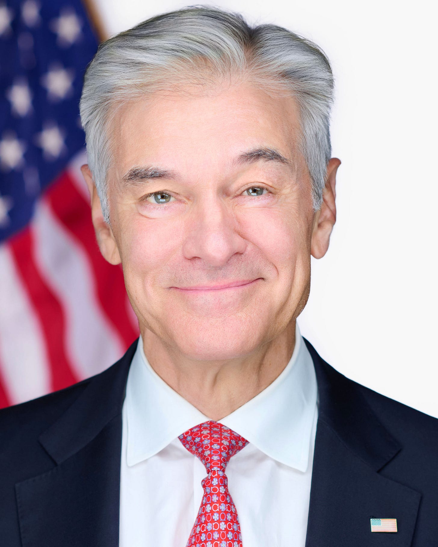 Dr. Mehmet Oz smiling in a dark suit and red patterned tie in front of an American flag