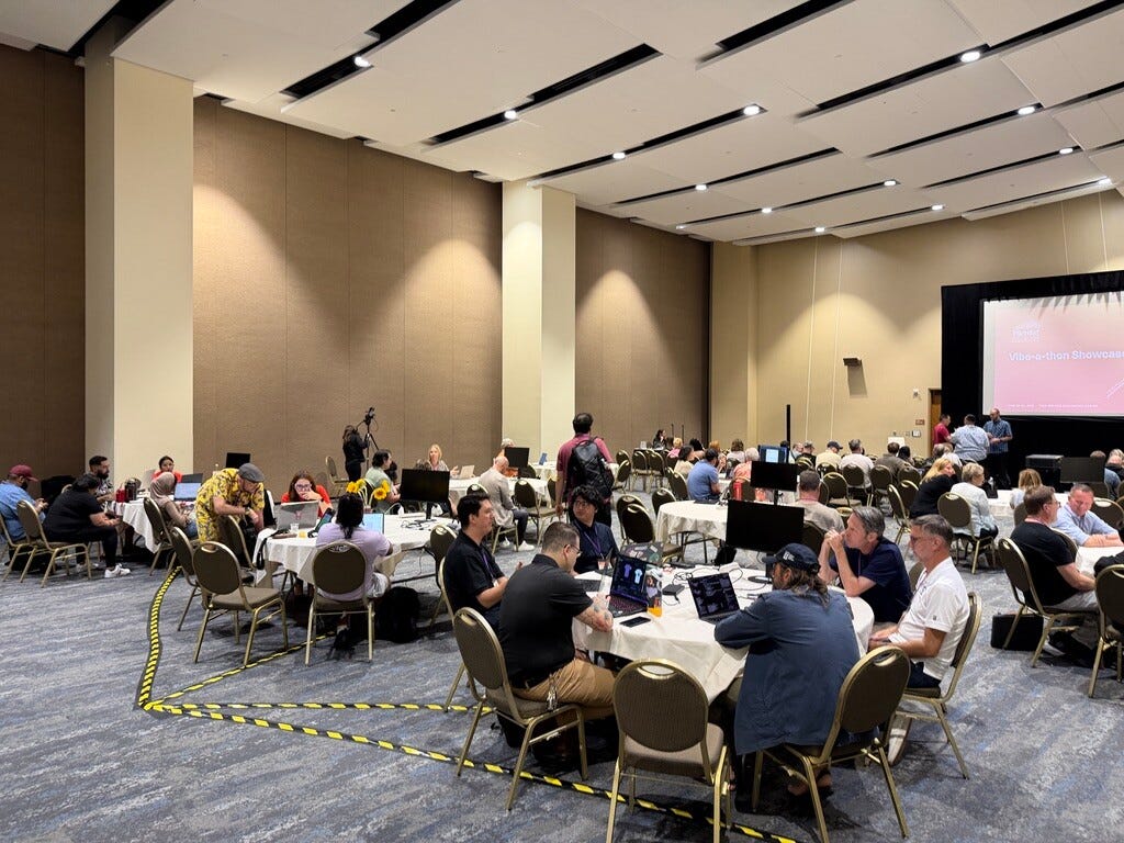 Attendees seated at round tables with laptops during a 'Vibe-a-thon Showcase' session, with a presentation screen visible in the background. Attendees seated at round tables with laptops during a 'Vibe-a-thon Showcase' session, with a presentation screen visible in the background.