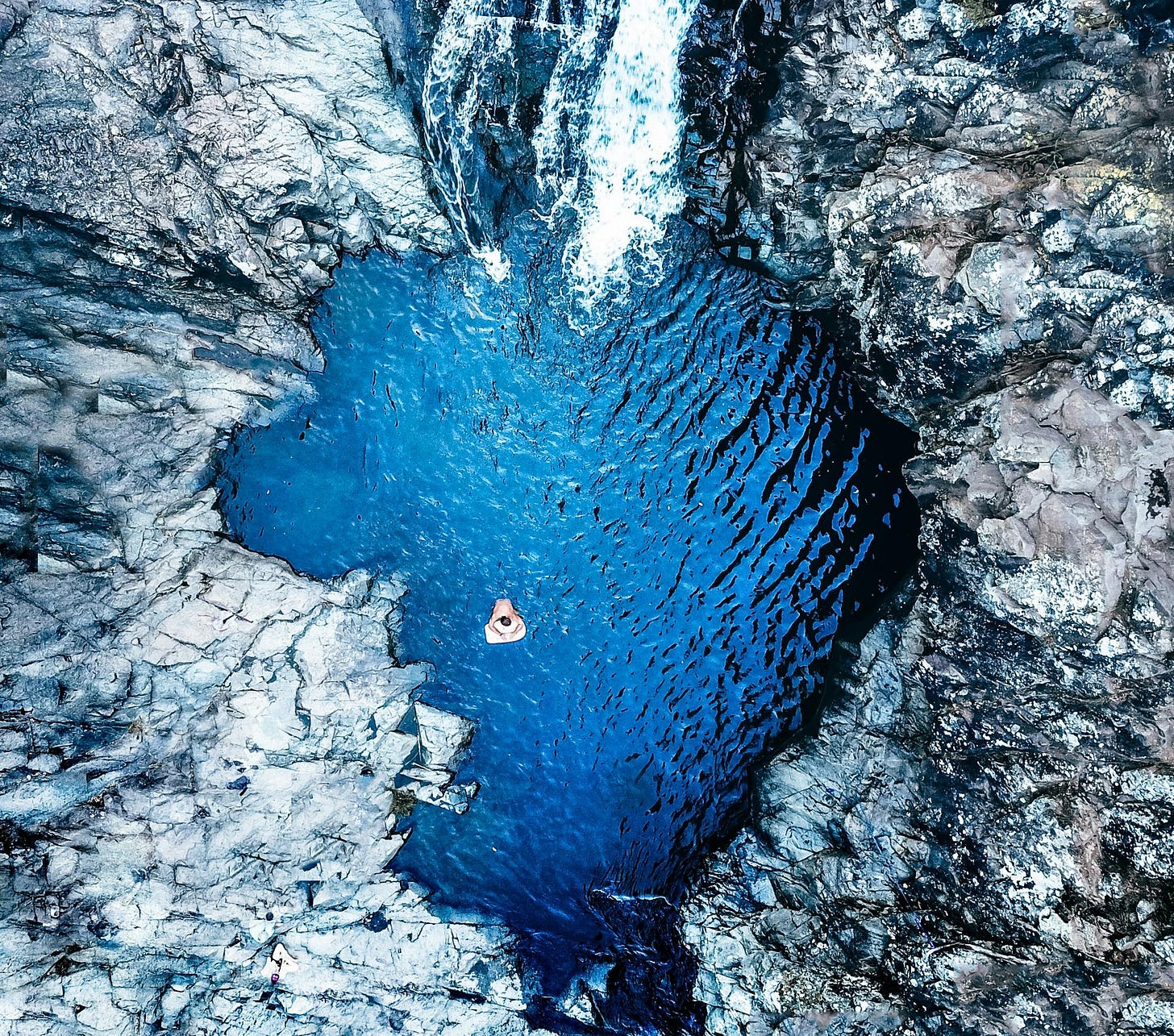 Aerial shot of a person immersed in a small, circular pool surrounded by rocky terrain. Water flows into the pool from a waterfall, creating a striking contrast between the deep blue water and rugged gray rocks. Aerial shot of a person immersed in a small, circular pool surrounded by rocky terrain. Water flows into the pool from a waterfall, creating a striking contrast between the deep blue water and rugged gray rocks.