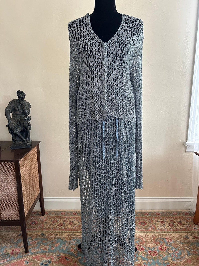 May include: A long, gray, crocheted dress with a V-neck and a single button closure. The dress has a loose fit and a flowing skirt. The dress is made of a lightweight, breathable fabric and is perfect for warm weather.