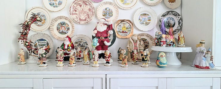 A festive collection of Santa figurines from various countries is displayed on a white cabinet. Decorative plates with scenic designs hang on the wall behind, enhancing the colorful and cheerful holiday atmosphere. A festive collection of Santa figurines from various countries is displayed on a white cabinet. Decorative plates with scenic designs hang on the wall behind, enhancing the colorful and cheerful holiday atmosphere.