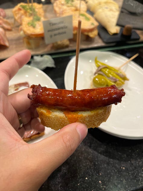 Crab stuffed squid, a spoon entering creamed uni in its shell, a hand holding a sesame crack with three different color pieces of smoked fish on top , a hand holding grilled foie on bread, a hand holding txistorra skewered onto a piece of bread, a fork entering a half eaten piece of cheesecake