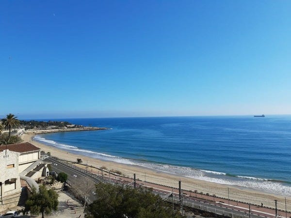 Vista do Balcão do Mediterrâneo, em Tarragona. Foto de Edmara Galvão. Descrição de Imagem: metade superior da imagem mostra um céu azul claro, sem nuvens. A parte inferior é, majoritariamente, um mar. Ele tem tons de azul escuro, com faixas mais claras. É um mar calmo. Com um barco ao fundo. Apesar de enorme, o barco está minúsculo. É possível ver a faixa de areia, uma ponta do litoral que se destaca para frente e uma parte da calçada projetada.