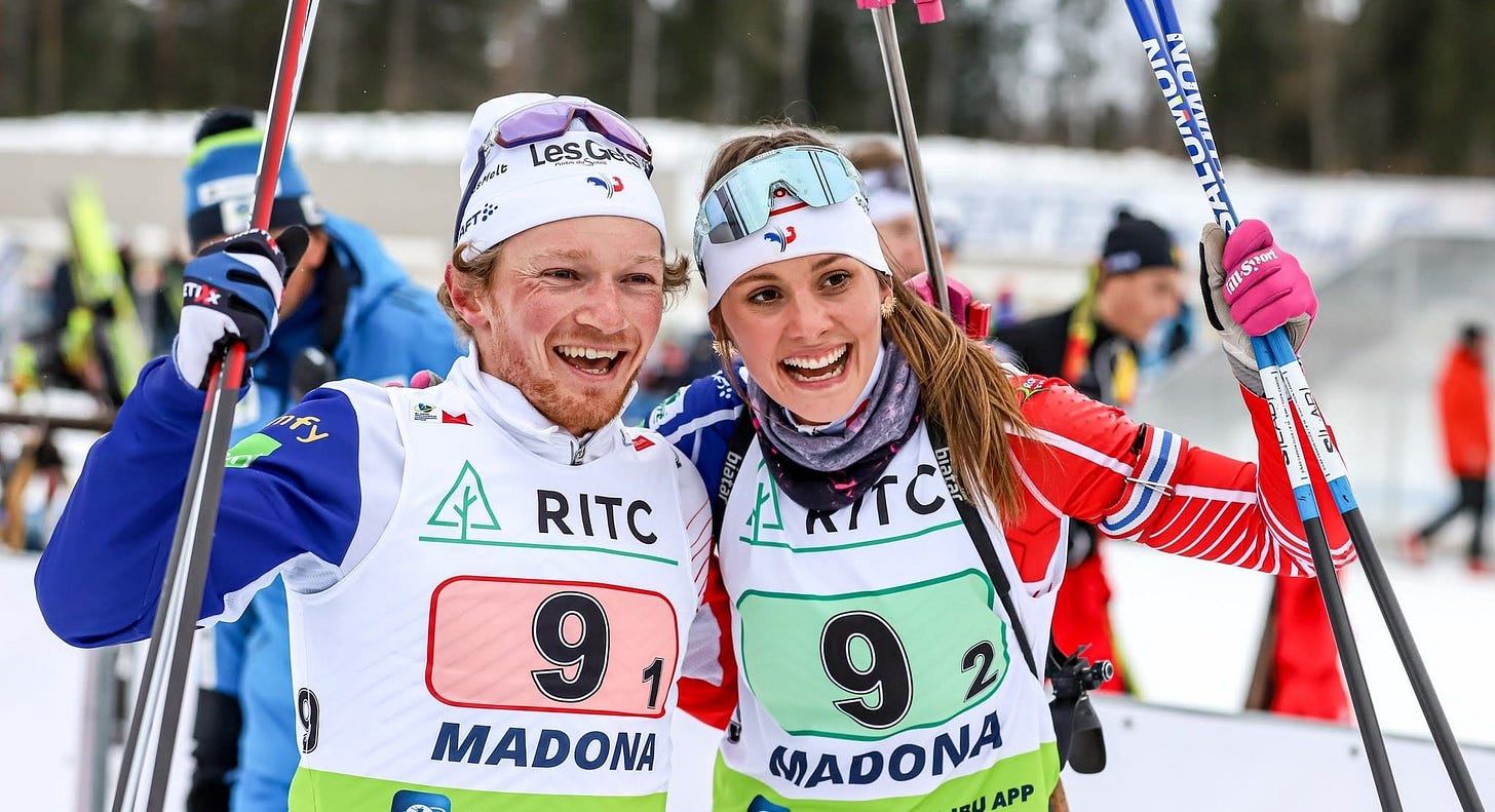 Two biathletes smiling and celebrating together at the finish line in Madona, Latvia, wearing race bibs number 91 and 92, arms around each other and holding ski poles in the air.