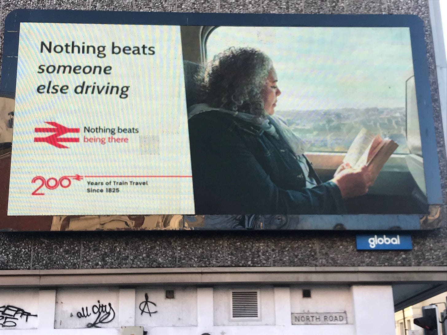 An electronic billboard showing a woman enjoying reading a book on a train. The caption reads, 'Nothing beats someone else driving; nothing beats being there.' There is the red symbol of National Rail (UK).